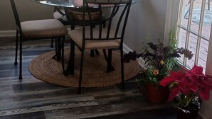 Un comedor con una mesa redonda, sillas y un jarrón con flores. Hay plantas en macetas y una ventana con cortinas.