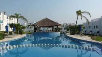 A swimming pool with a thatched-roof hut in the center, surrounded by white buildings and palm trees.