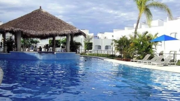 A swimming pool with a thatched-roof structure, palm trees, and white buildings in the background. - Departamento en Tequesquitengo (Tequesquitengo)