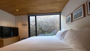 A bedroom with a large bed, a TV on a wooden dresser, and a stone wall visible through a large window.