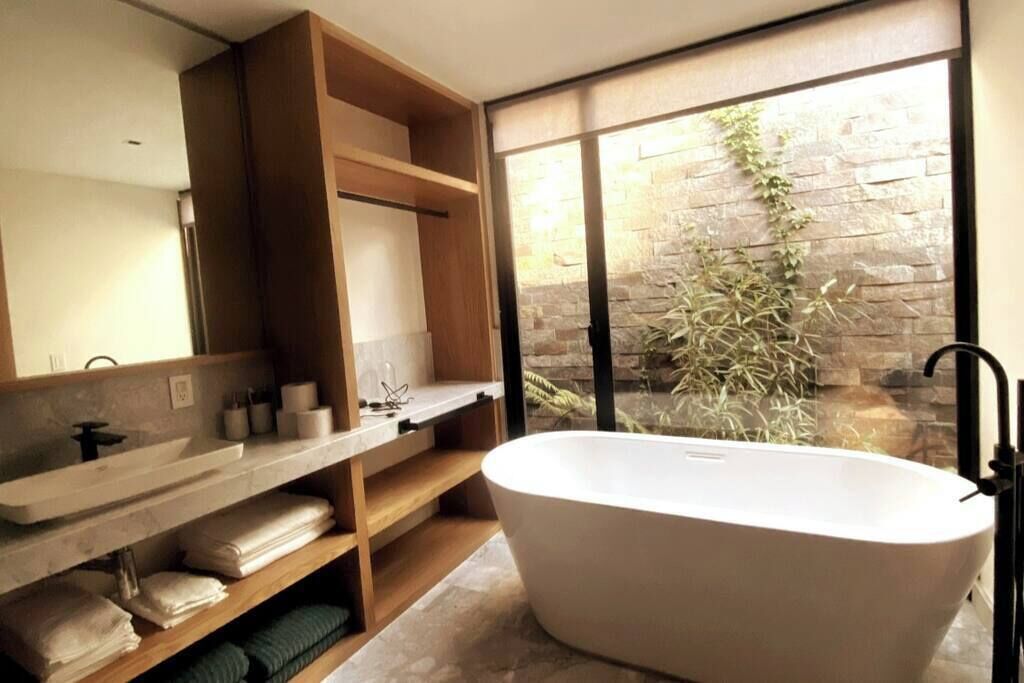 A modern bathroom with a freestanding bathtub, wooden shelves, a large mirror, and a stone wall with greenery.