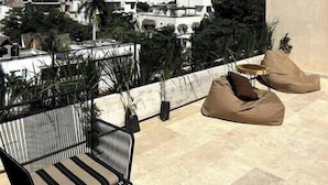 Eine Dachterrasse mit Blick auf eine Stadtlandschaft, darunter weiße Gebäude und Grünflächen. Zwei beige Sitzsäcke und ein schwarzer Metallstuhl mit Streifenmuster sind vorhanden.