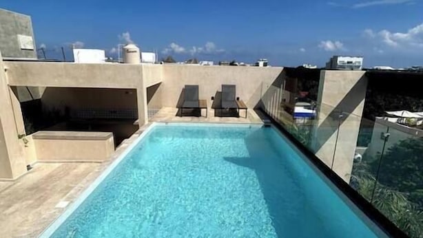 Ein Pool auf dem Dach mit klarem, blauem Wasser, zwei Liegestühle und Blick auf die Skyline der Stadt unter einem bewölkten Himmel.
