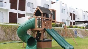 A wooden play structure with a slide and climbing area, set against a backdrop of modern residential buildings. - Depto con Alberca y Vista al Lago en Teques (Tequesquitengo)