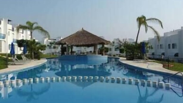 A swimming pool with a thatched-roof structure in the center, surrounded by white buildings and palm trees. - Depto con Alberca y Vista al Lago en Teques (Tequesquitengo)
