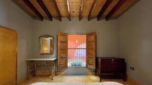 A bedroom with a wooden ceiling, a bed, a dresser, a mirror, and a window with shutters.