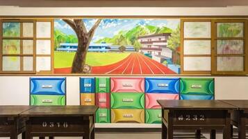 Une pièce ornée d’une grande fresque représentant un chemin bordé d’arbres menant à un bâtiment, ainsi que de rangements colorés et d’une table avec des tiroirs.