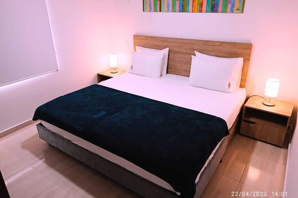 A neatly made bed with a dark blue blanket, two white pillows, and a wooden headboard. There are two bedside tables with lamps, and a colorful abstract painting on the wall.