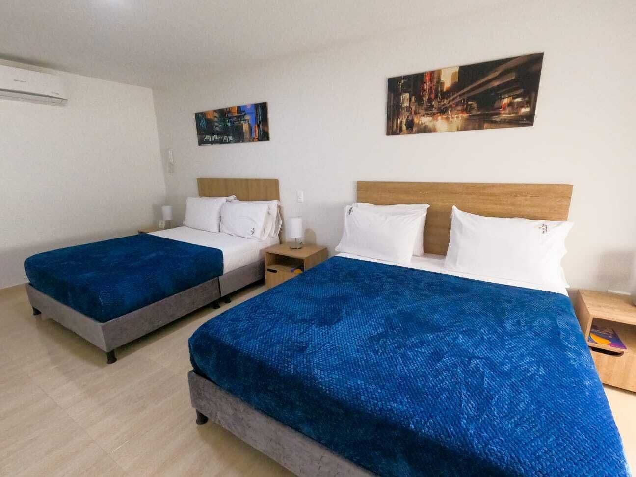 A hotel room with two single beds and one double bed, each with a blue bedspread. There are two nightstands with lamps, and the walls are adorned with two framed pictures.