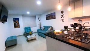 Ein kompakter Wohnbereich mit Sofa, Hocker und Couchtisch. Ein Küchenbereich mit Herd und Spüle ist sichtbar.