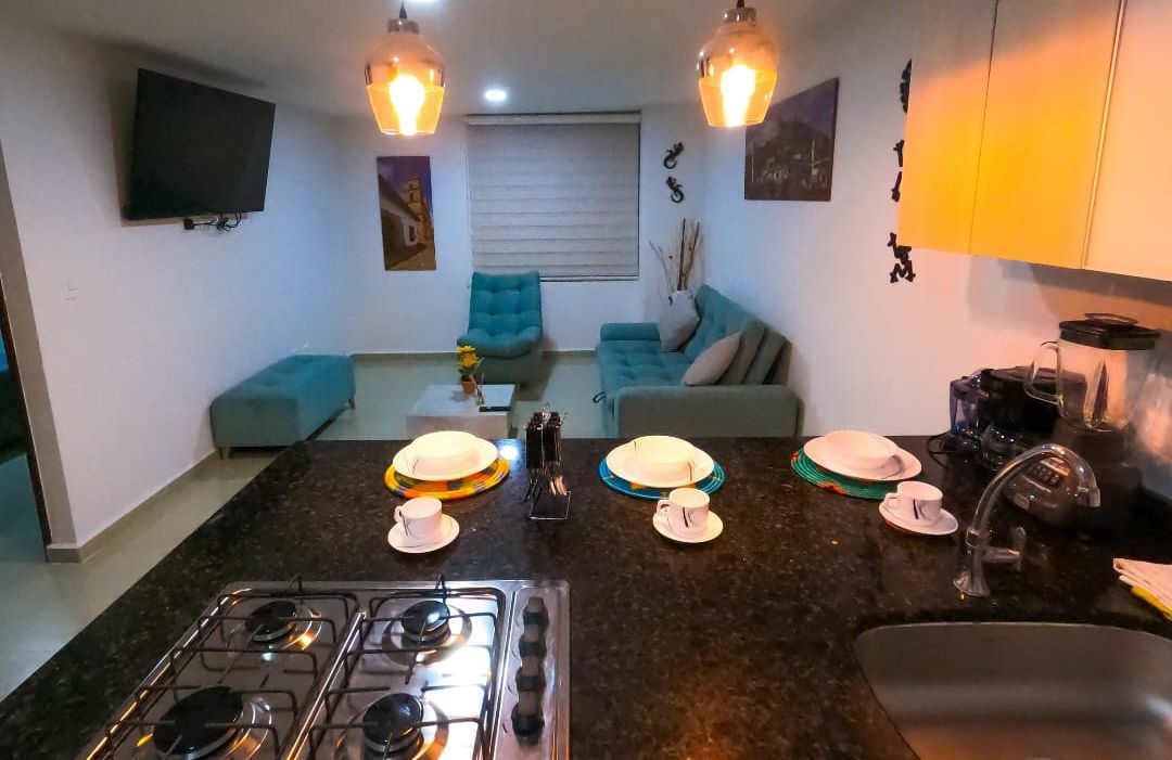 Cocina compacta con estufa a gas, un lavamanos y una zona de comedor con una mesa para dos. Hay un sofá y un televisor en la sala.