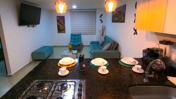 Cocina compacta con estufa a gas, un lavamanos y una zona de comedor con una mesa para dos. Hay un sofá y un televisor en la sala.