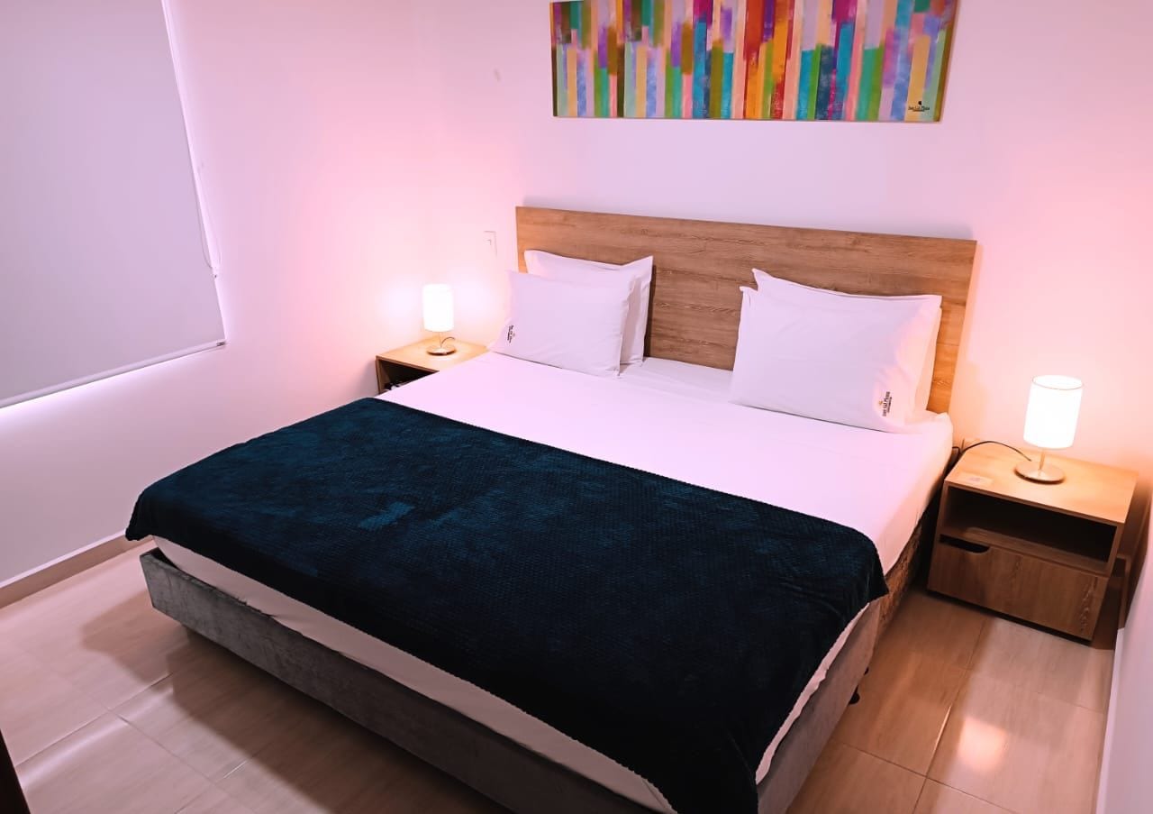 Una cama bien hecha con una colcha azul oscuro, dos almohadas blancas y un cabecero de madera. Hay dos mesitas de noche con lámparas y un cuadro abstracto colorido en la pared.