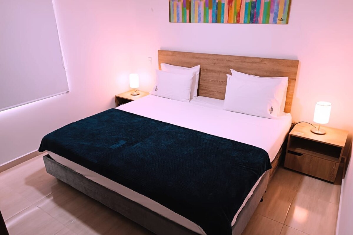 Una cama bien hecha con una colcha azul oscuro, dos almohadas blancas y un cabecero de madera. Hay dos mesitas de noche con lámparas y un cuadro abstracto colorido en la pared.