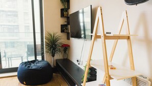 Un salon moderne avec une grande fenêtre, un fauteuil poire noir, un téléviseur à écran plat, un chevalet en bois et une petite bibliothèque.