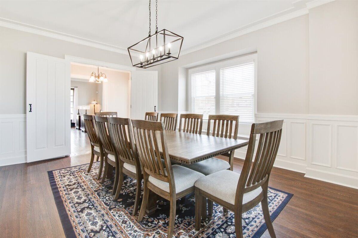 A dining room with a rectangular wooden table surrounded by six chairs, a patterned rug, and a hanging light fixture.