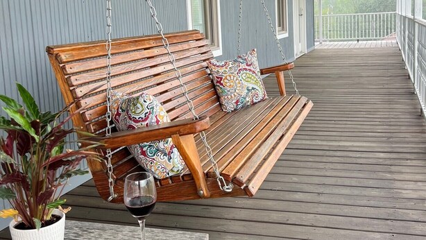 Een houten veranda met een schommelzitje, een tafel met een glas wijn en een bloempot.