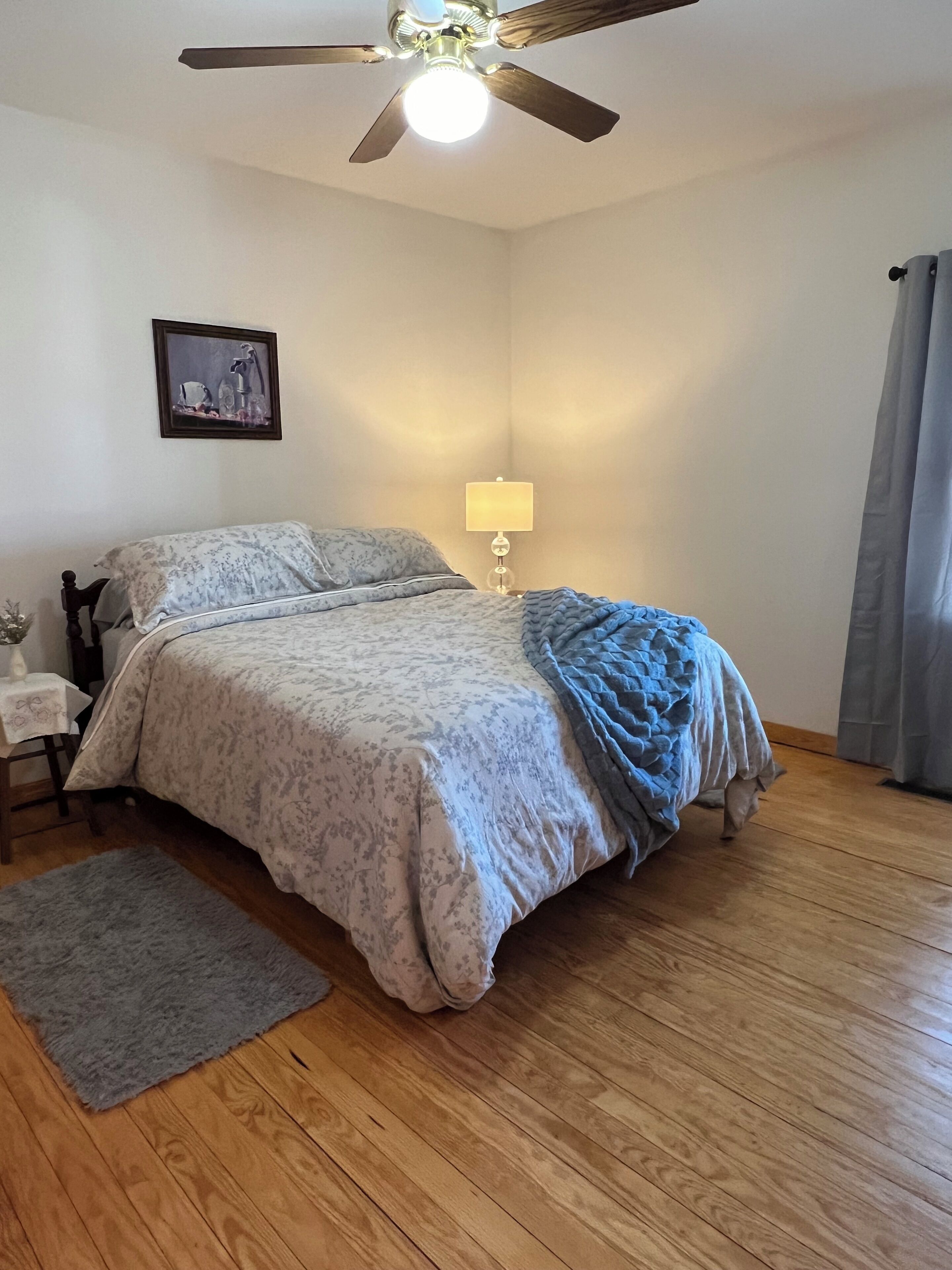 Une chambre à coucher avec un lit, une table de chevet, une lampe, un ventilateur de plafond, un tableau au mur et une fenêtre avec des rideaux.