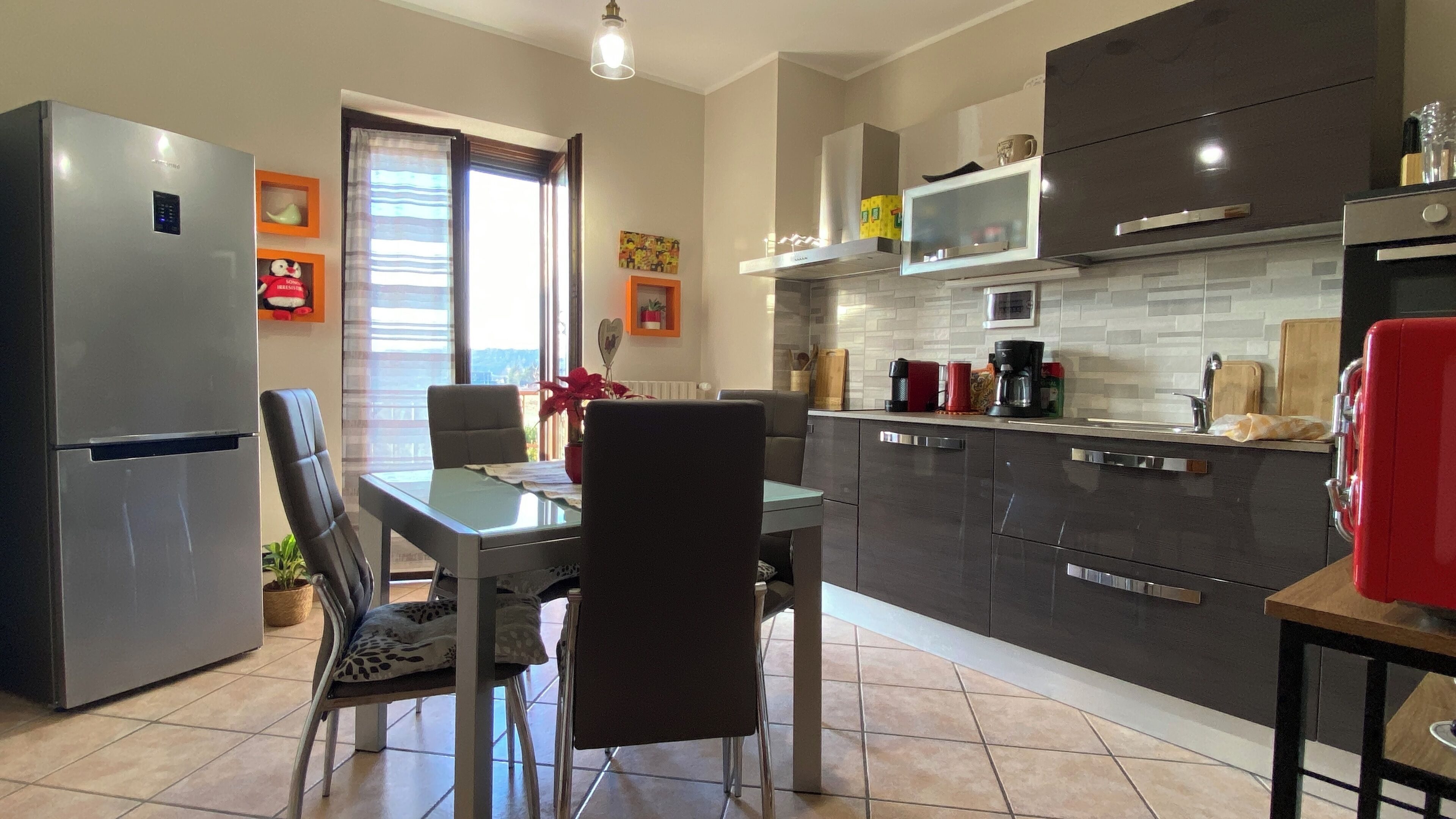 A modern kitchen with dark cabinetry, a stainless steel refrigerator, a dining table with chairs, and a window with curtains.