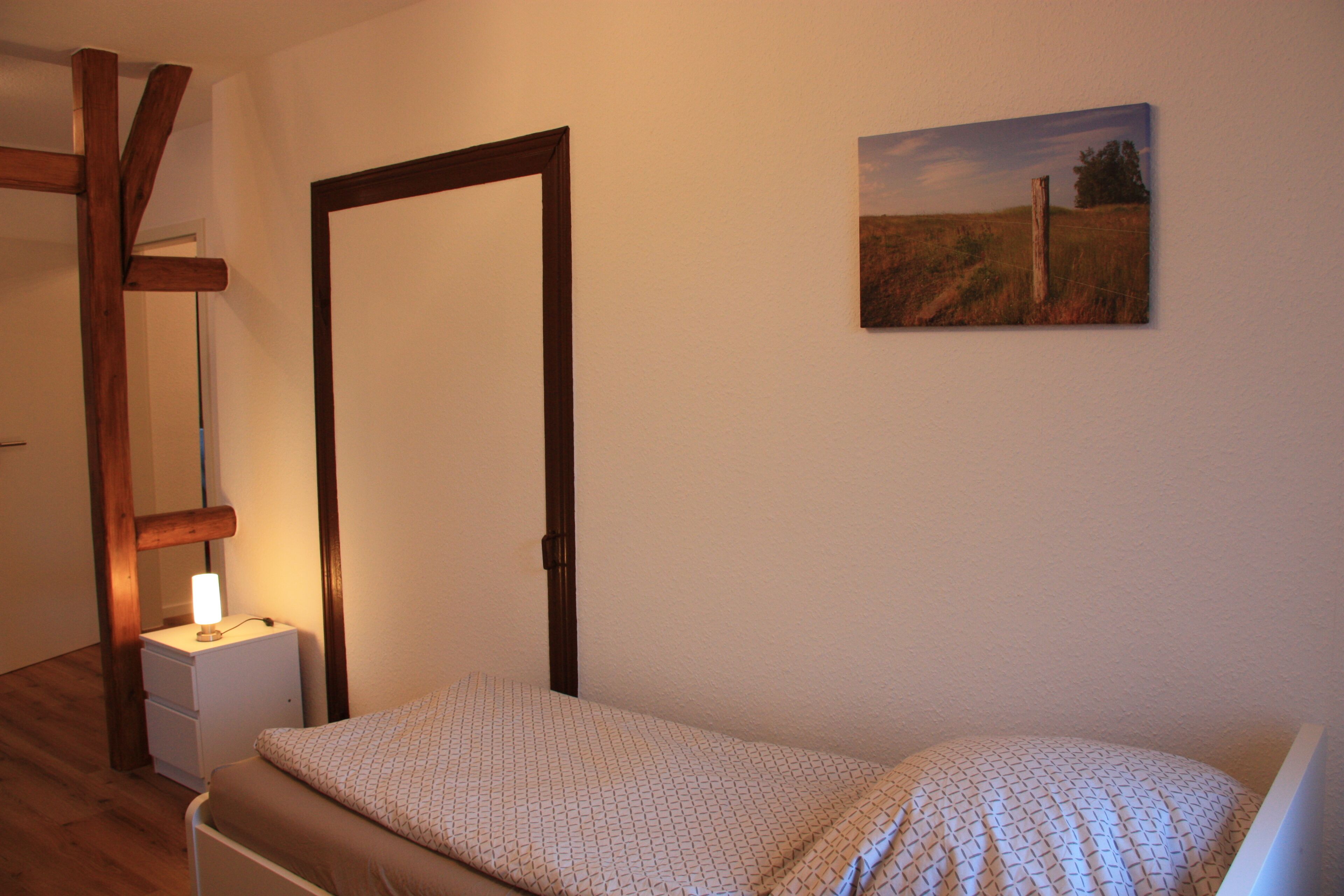 A bedroom with a bed, a nightstand, a lamp, a door, and a painting on the wall.