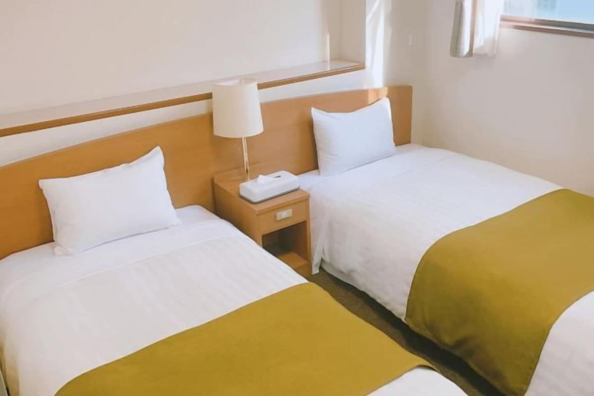 A hotel room with two beds, a nightstand, a lamp, and a window with curtains.