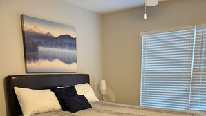 A bedroom with a bed, a nightstand, a lamp, a ceiling fan, a window with blinds, and a painting on the wall. - Lake Hartwell/Clemson University 3 bd/2 bath condo (Anderson)