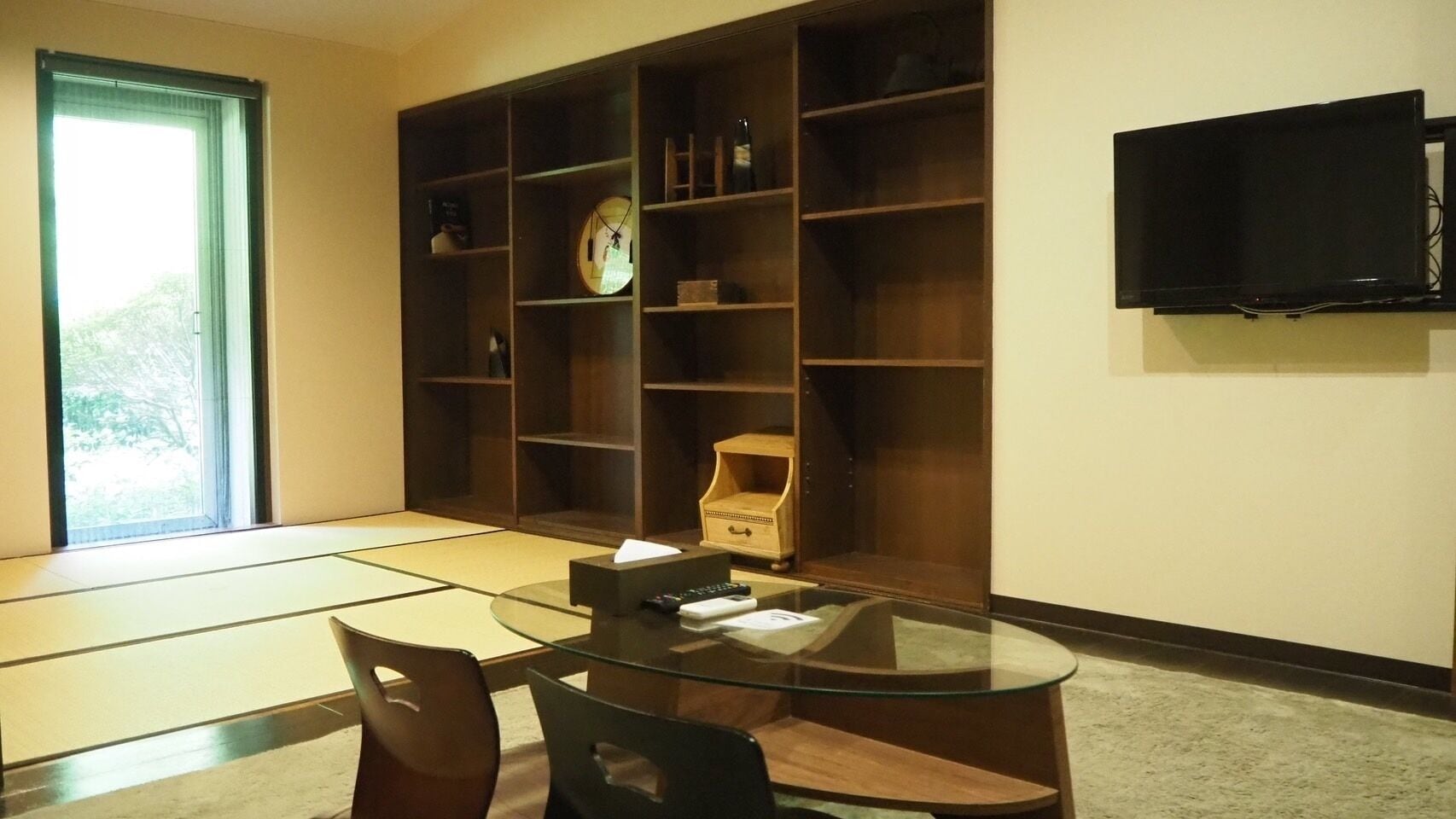 A room with tatami mats, a glass table, chairs, a wooden cabinet, a clock, and a TV on the wall.