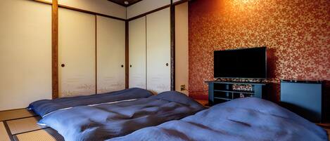 A traditional Japanese room with tatami flooring, two tatami mats, a television on a stand, and a wooden ceiling.