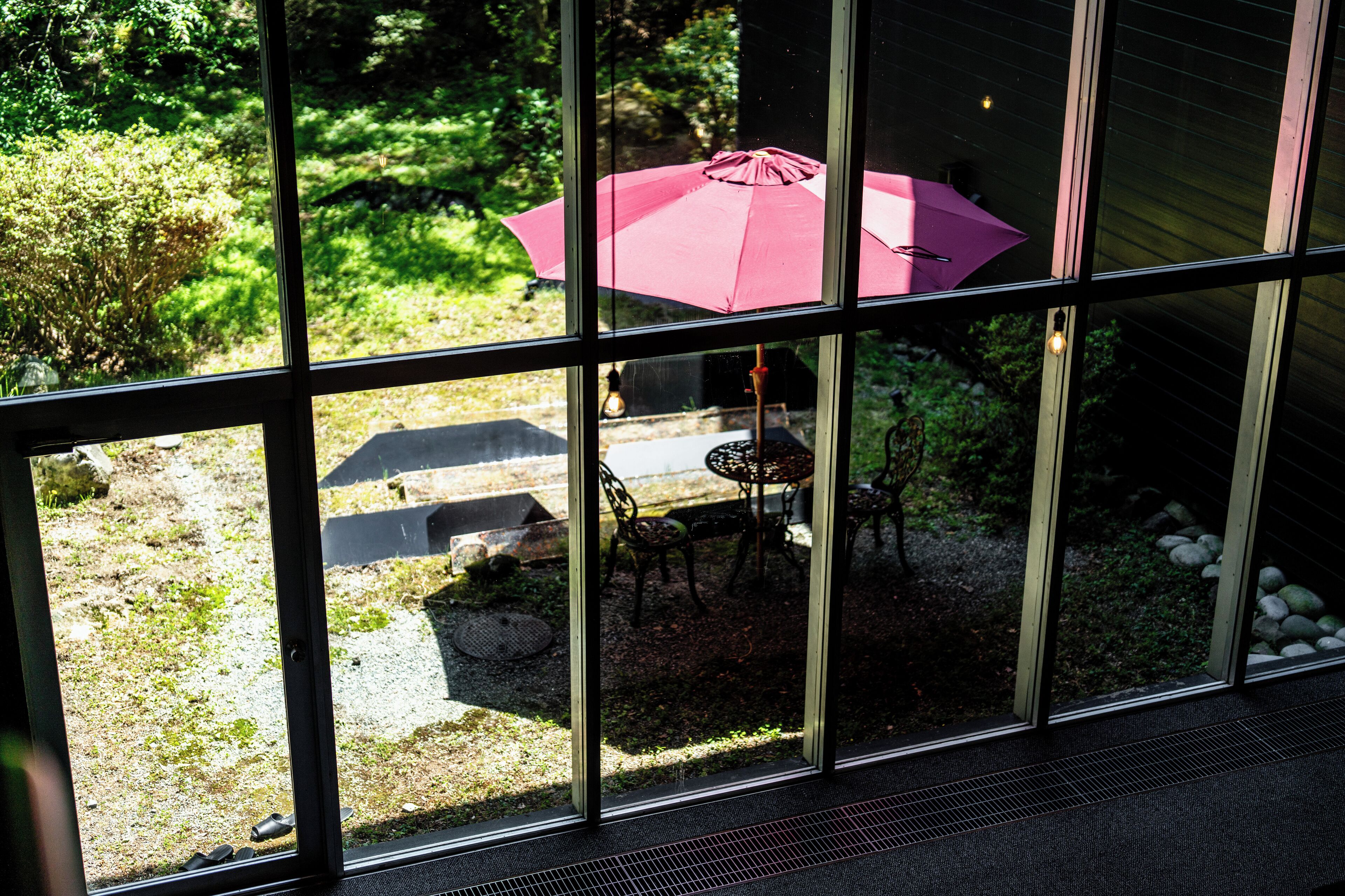 Blick durch ein Fenster auf eine Terrasse mit einem roten Sonnenschirm, Gartenmöbeln und einem gepflasterten Weg.
