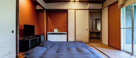 Et traditionelt japansk værelse med tatamigulv, en seng med blåt sengetøj, et fjernsyn og en aircondition.