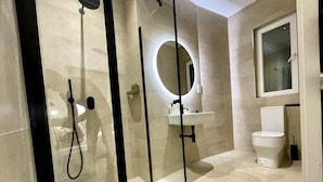 Un baño moderno con ducha de vidrio, un espejo redondo, inodoro y lavamanos.