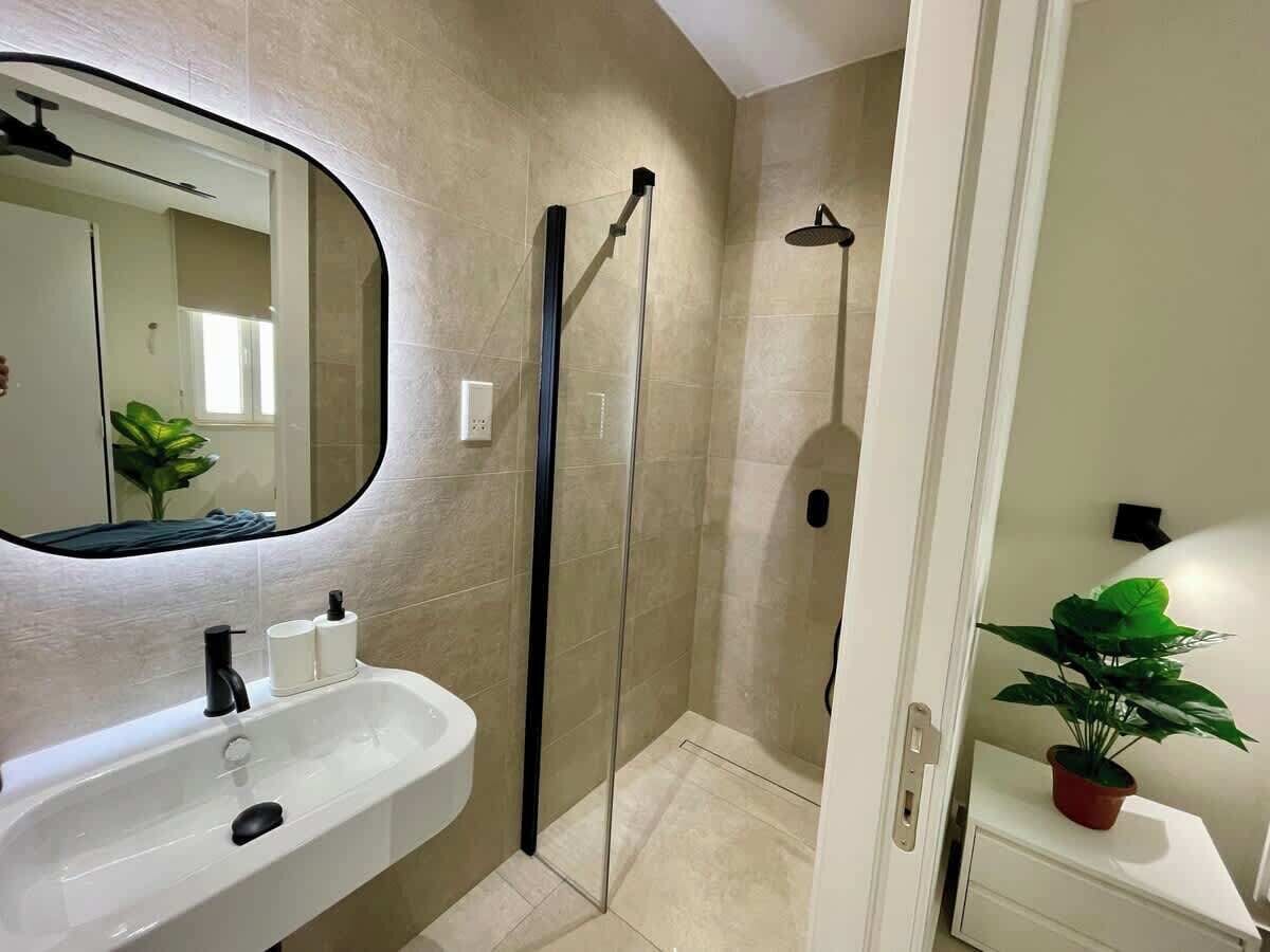 Un baño con lavamanos blanco, espejo, cabina de ducha y planta en maceta.