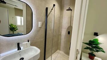 Un baño con lavamanos blanco, espejo, cabina de ducha y planta en maceta.