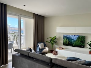 A modern living room with a sofa, armchair, TV, and a balcony with a view.