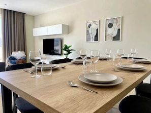 Une table à manger dressée avec des assiettes, des verres et des couverts, dans une pièce moderne agrémentée d’un téléviseur, d’œuvres d’art et d’une plante.