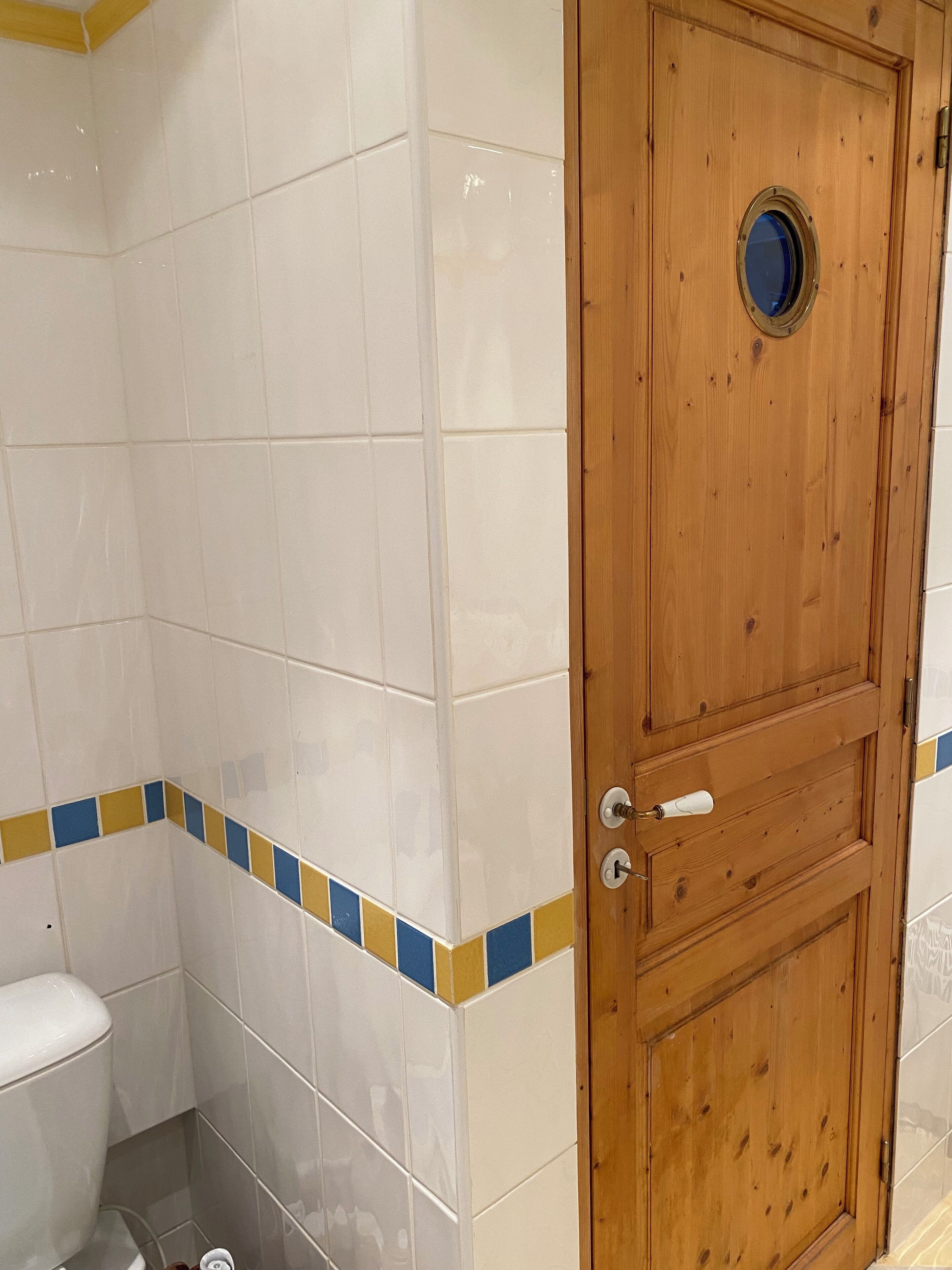 Un bagno con piastrelle bianche, una porta in legno con un oblò tondo e un angolo decorato con piastrelle azzurre e gialle.
