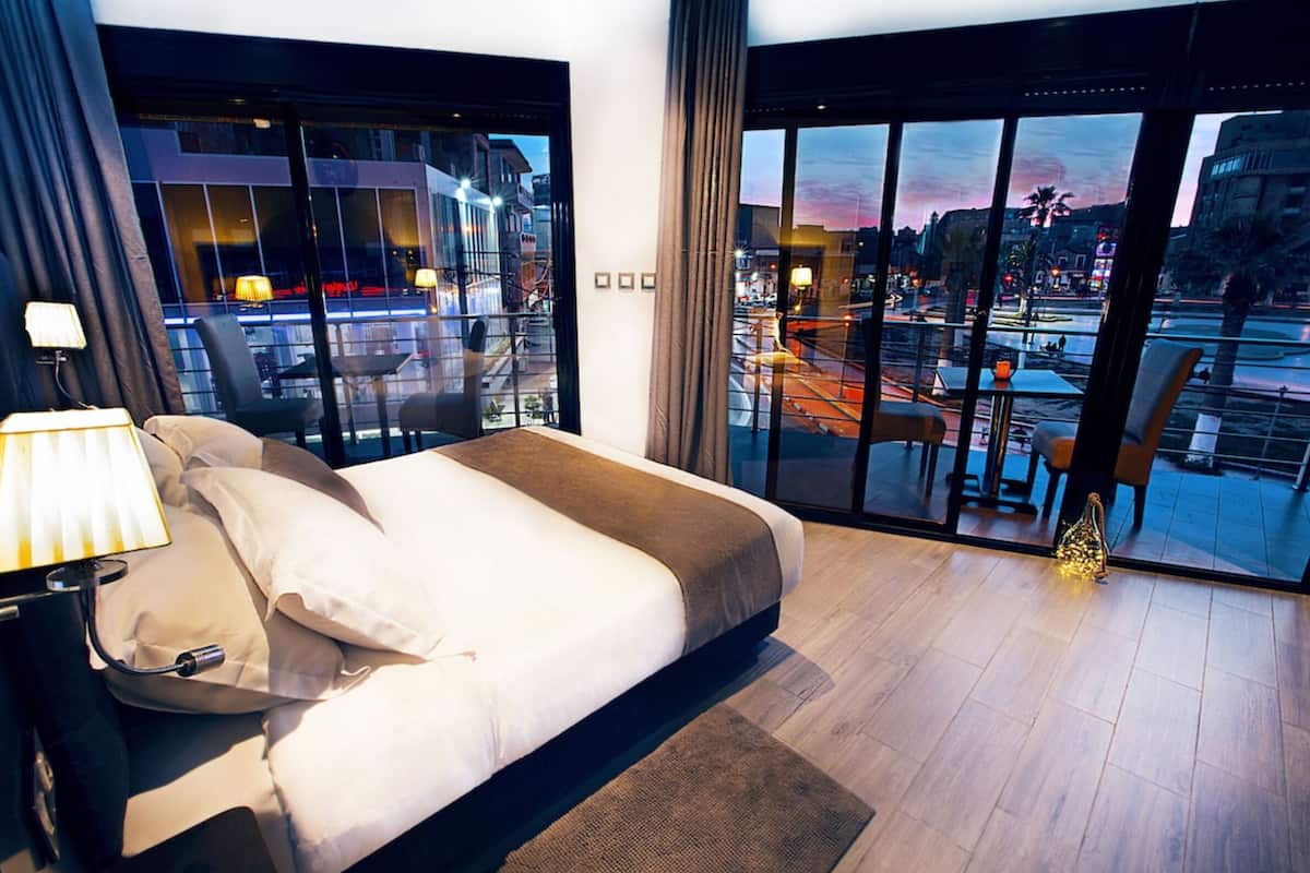 A modern hotel room with a large bed, a nightstand, and a view of a cityscape and pool area through floor-to-ceiling windows.