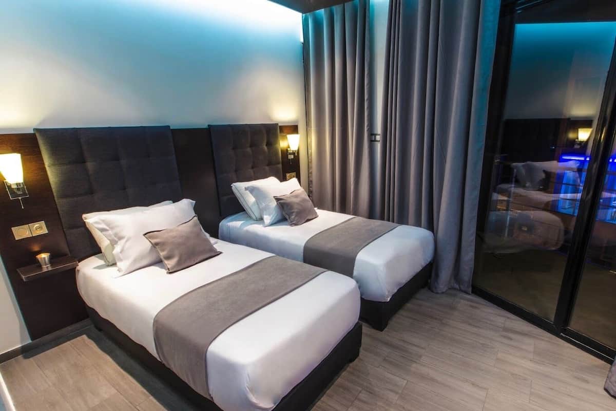 A hotel room with two beds, a dark headboard, a nightstand, and a large window with curtains.