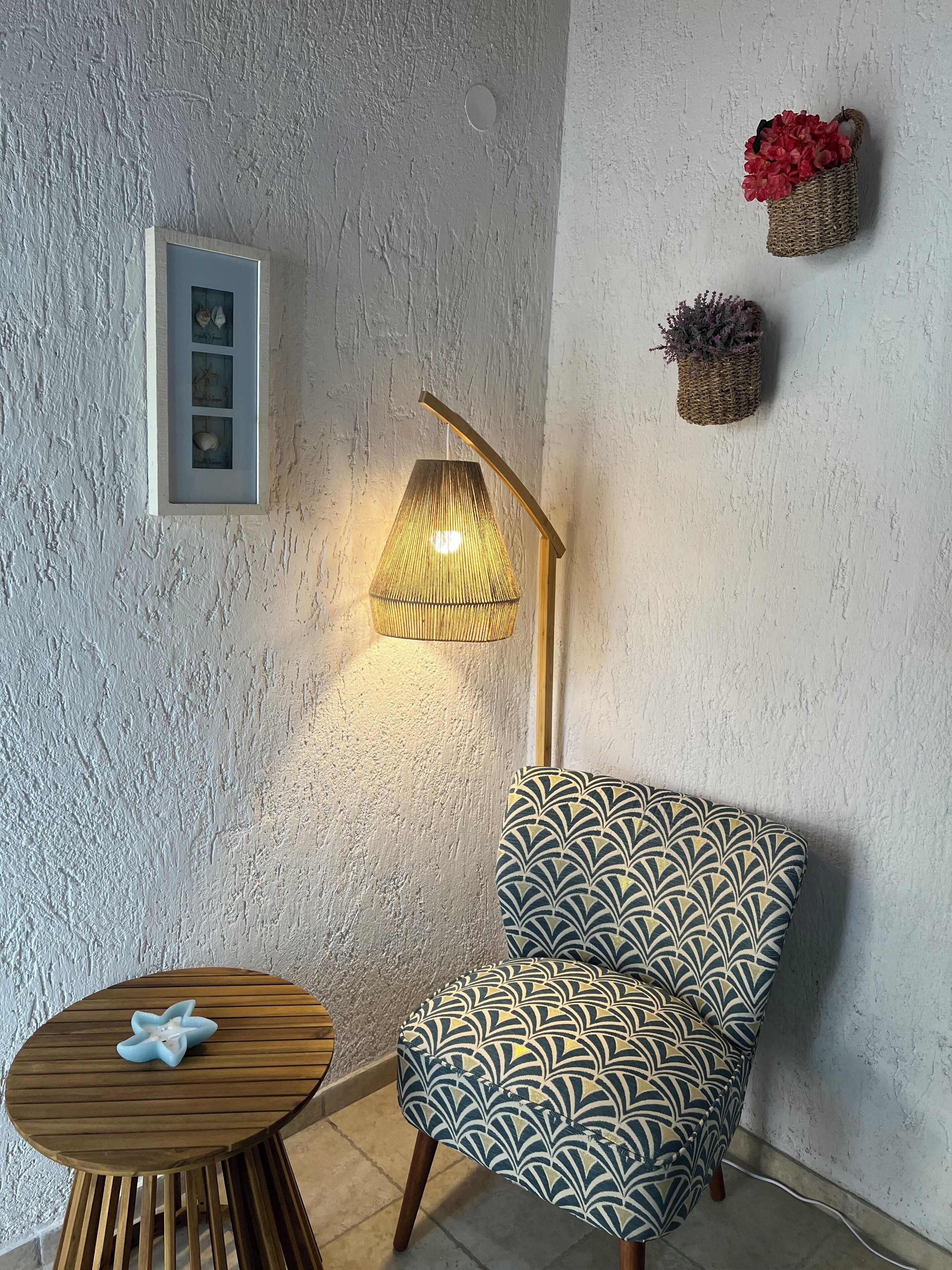 A room with a textured white wall, a wooden side table with a blue object, a patterned armchair, a floor lamp, and two wall-mounted baskets with flowers.