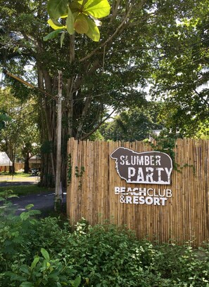 Señal de 'Sumber Party Beach Club & Resort' con valla de bambú y exuberante follaje.