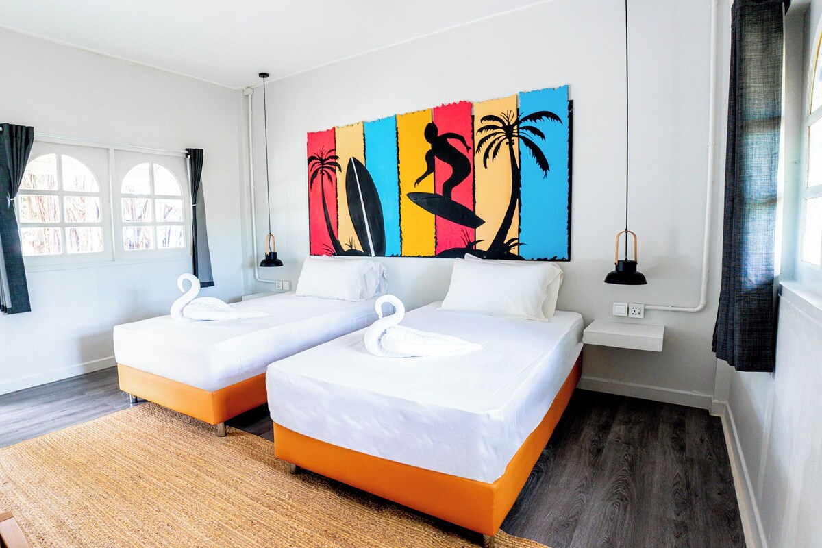 A hotel room with two beds, a large colorful artwork on the wall, a window with curtains, and a small table with a lamp.