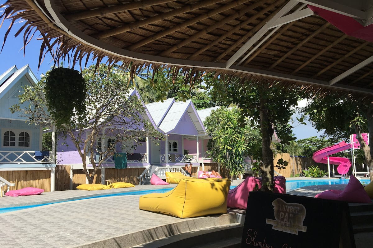 Un espace piscine avec des fauteuils en forme de sac de haricots jaunes, un panneau indiquant les prix des « Slumber Shakes » (12 dollars) et un toboggan rose.