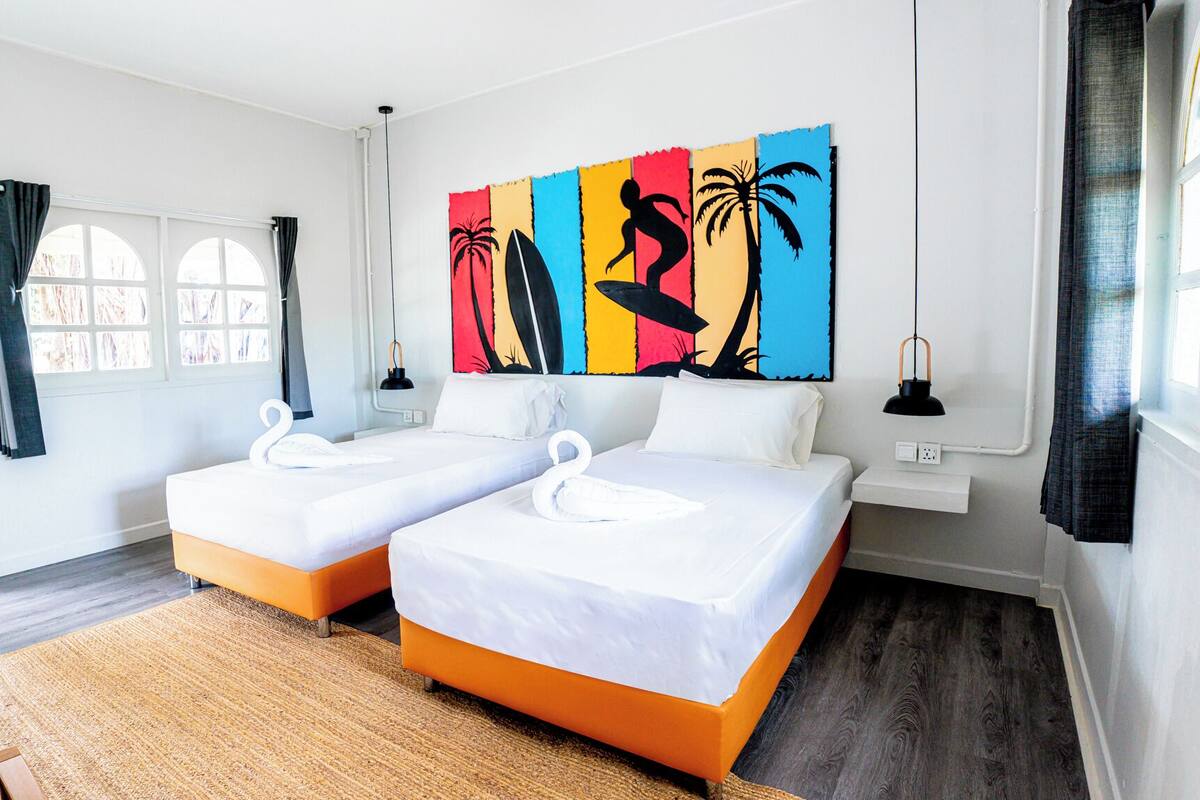 Une chambre d’hôtel avec deux lits, une grande œuvre d’art colorée accrochée au mur, une fenêtre avec des rideaux et une petite table avec une lampe.