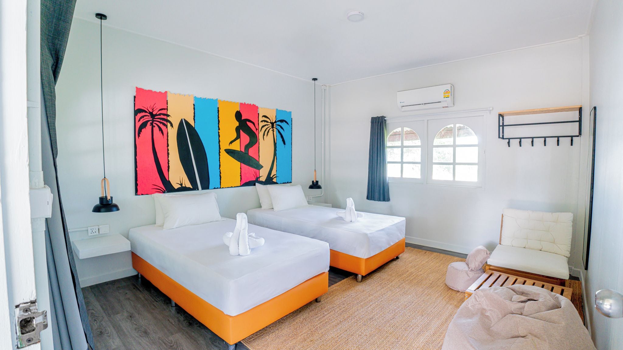 A hotel room with two beds, a large beach-themed artwork, a window with curtains, and an air conditioning unit.