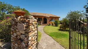 A stone wall with a sign, a pathway leading to a house with a tiled roof, and a metal gate.