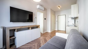 Ein modernes Wohnzimmer mit Flachbildfernseher, integriertem Stauraum, einem grauen Sofa und einer Küchenzeile mit Herd und Schränken.