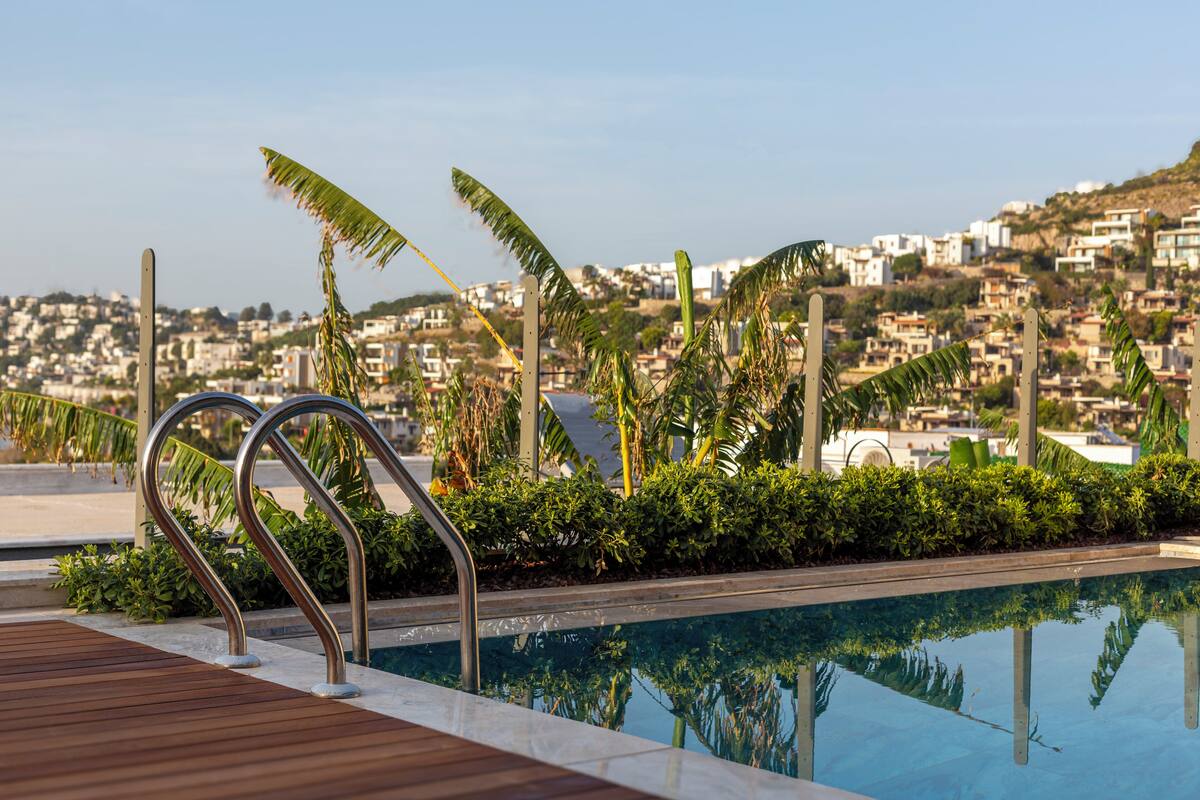 A swimming pool with metal railings, surrounded by greenery and overlooking a hillside residential area.