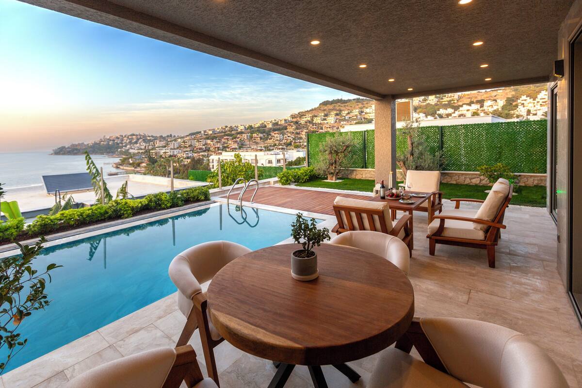 A poolside area with a wooden table and chairs, a potted plant, and a view of the ocean and hillside town.