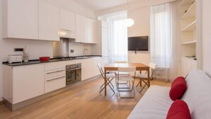 A modern kitchen with white cabinets, a dining area with a wooden table and chairs, and a living area with a sofa and a TV. - Testaccio Elegant Apartment (Rome)