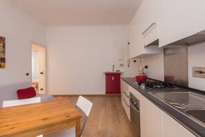 Een compacte keuken met een fornuis, spoelbak en kastjes. Er is een eethoek met een houten tafel en stoelen.